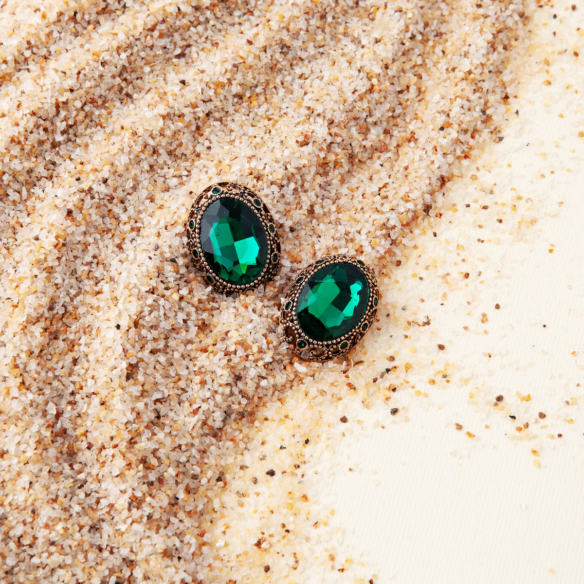 Pair of oval emerald green gem buttons with antique gold filigree settings half buried in sand