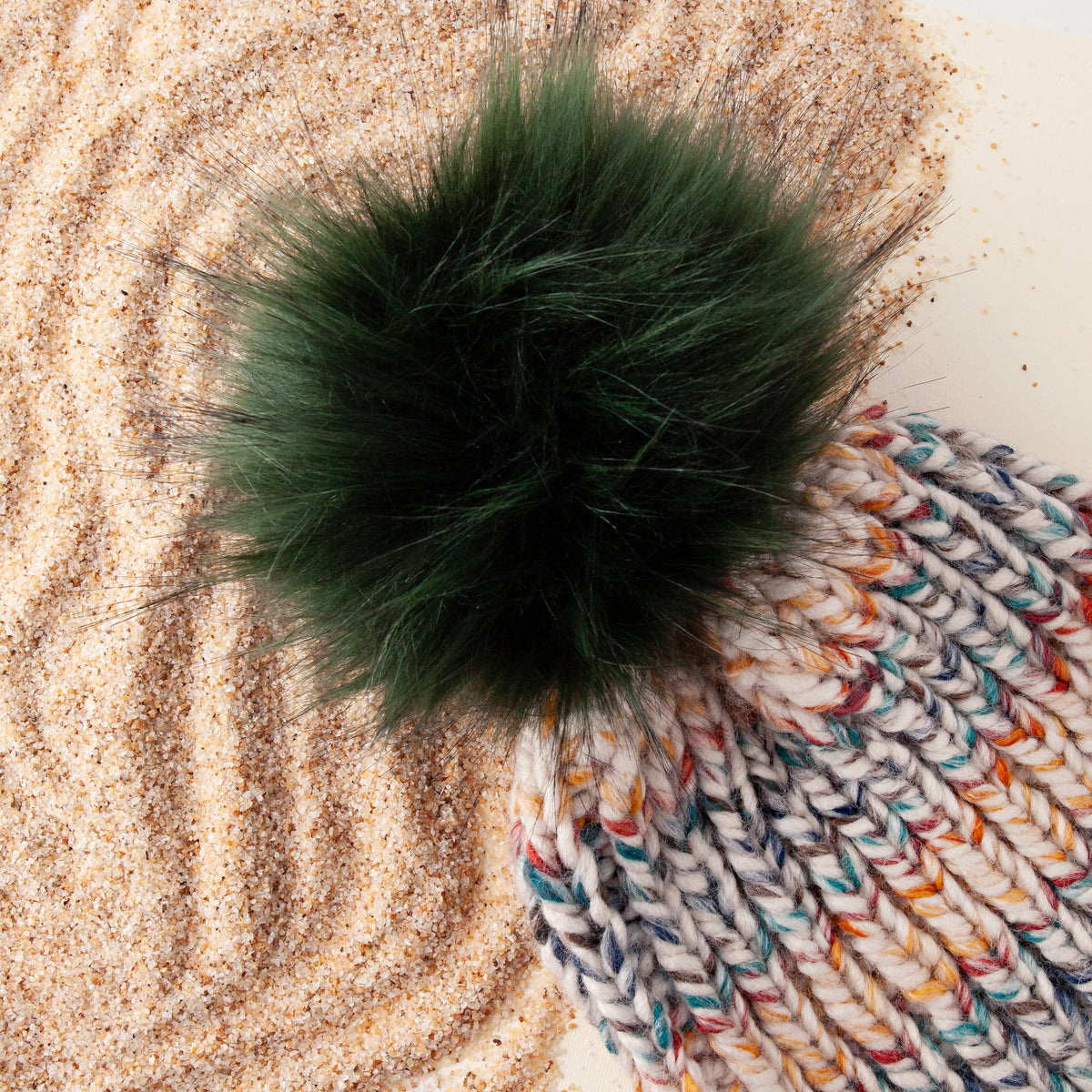 Forest green faux fur pom pom on a multicolor chunky knit hat resting on raked sand