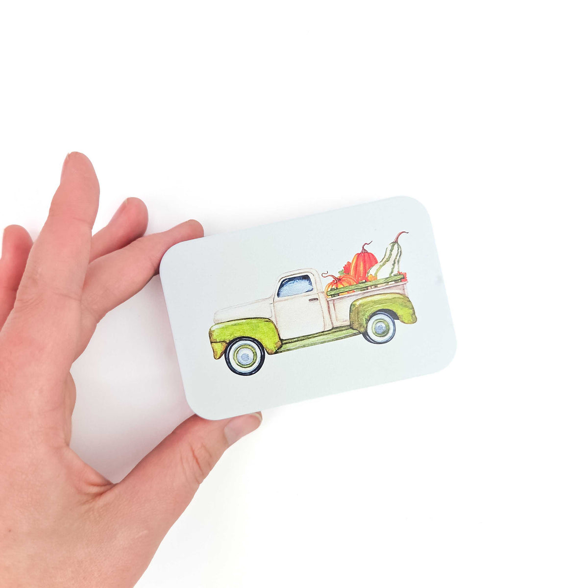 Hand holding the Pumpkin Truck stitch marker storage tin to show its palm-sized scale, lid painted with a watercolor pumpkin-filled truck