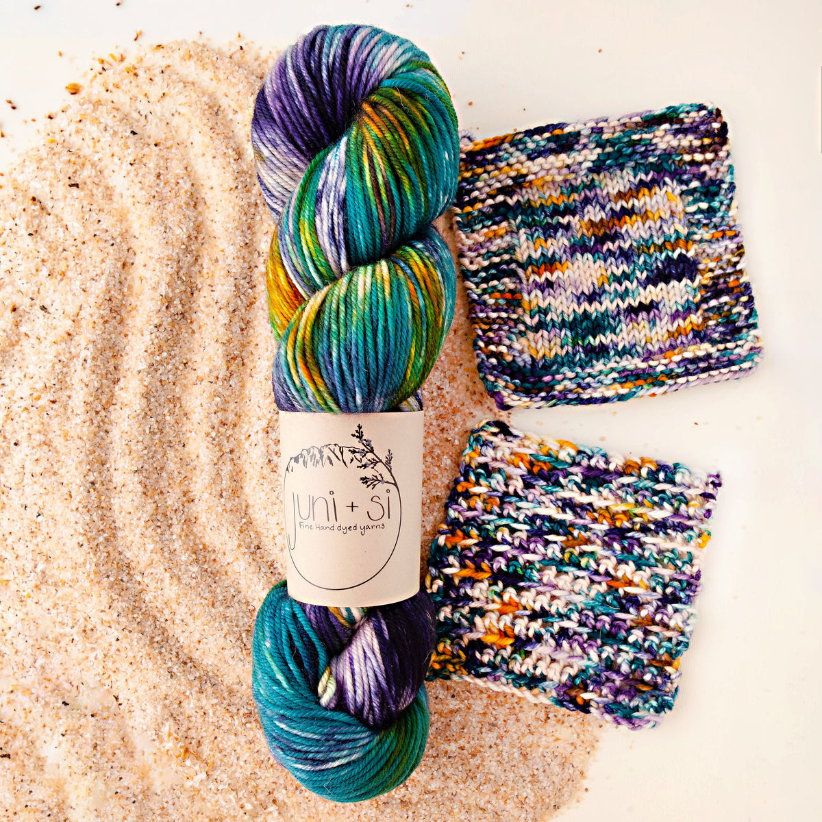 Two skeins of Juni + Si hand dyed yarn in teal, purple, green and gold with matching knit and crochet swatches on sand