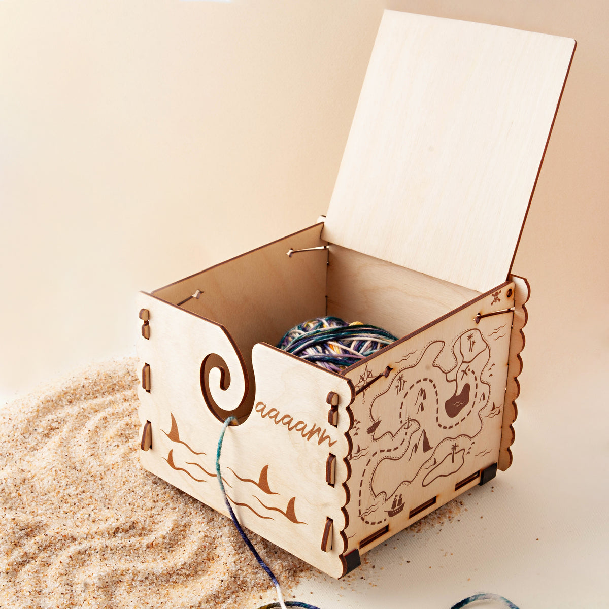 Laser-cut wooden yarn box with an engraved treasure map, shark fins and Yaaaarrr lettering, lid open with a yarn cake inside