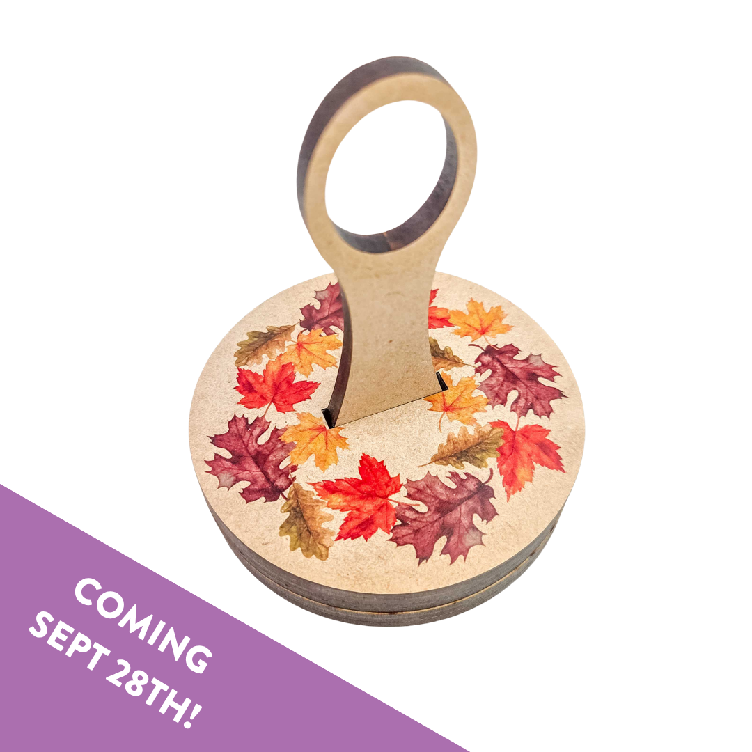 Round wooden carousel yarn holder printed with a wreath of red, orange and gold fall leaves, no wrist strap, with a Coming Sept 28th banner