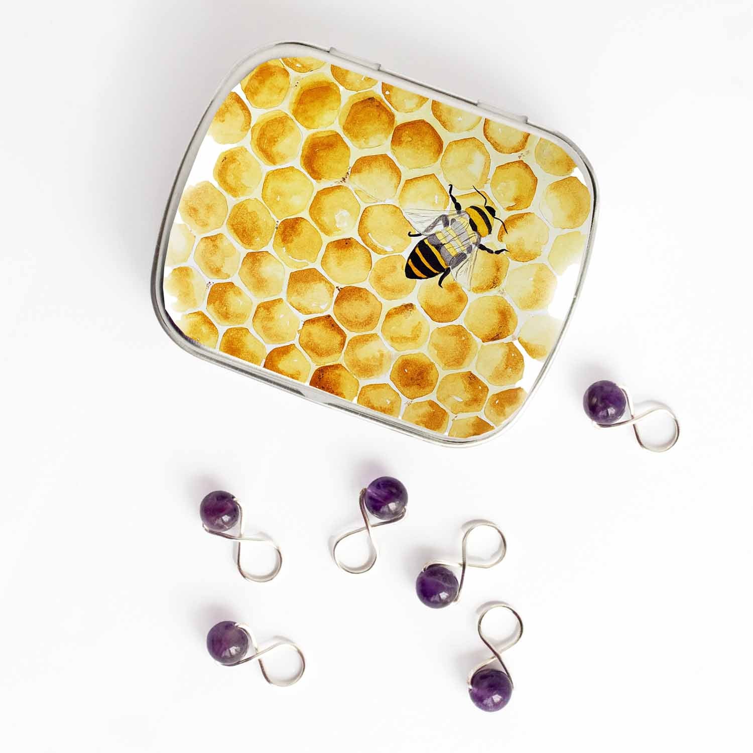 Small hinged Honey Bee tin with watercolor honeycomb lid, shown beside six purple beaded stitch markers