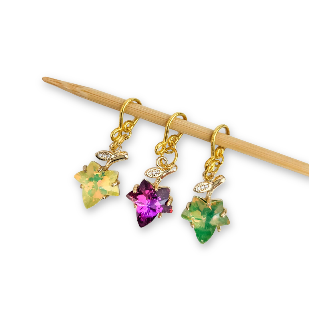 Three gold Fall Leaves Melody snag free stitch markers in yellow, magenta, and green crystal maple leaf shapes hanging from a bamboo knitting needle