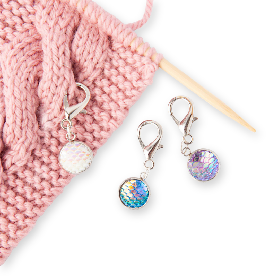 Three iridescent mermaid scale stitch markers with lobster clasp closures in white, teal, and purple on pink knitted fabric from the 2026 Mermaid Summer Box