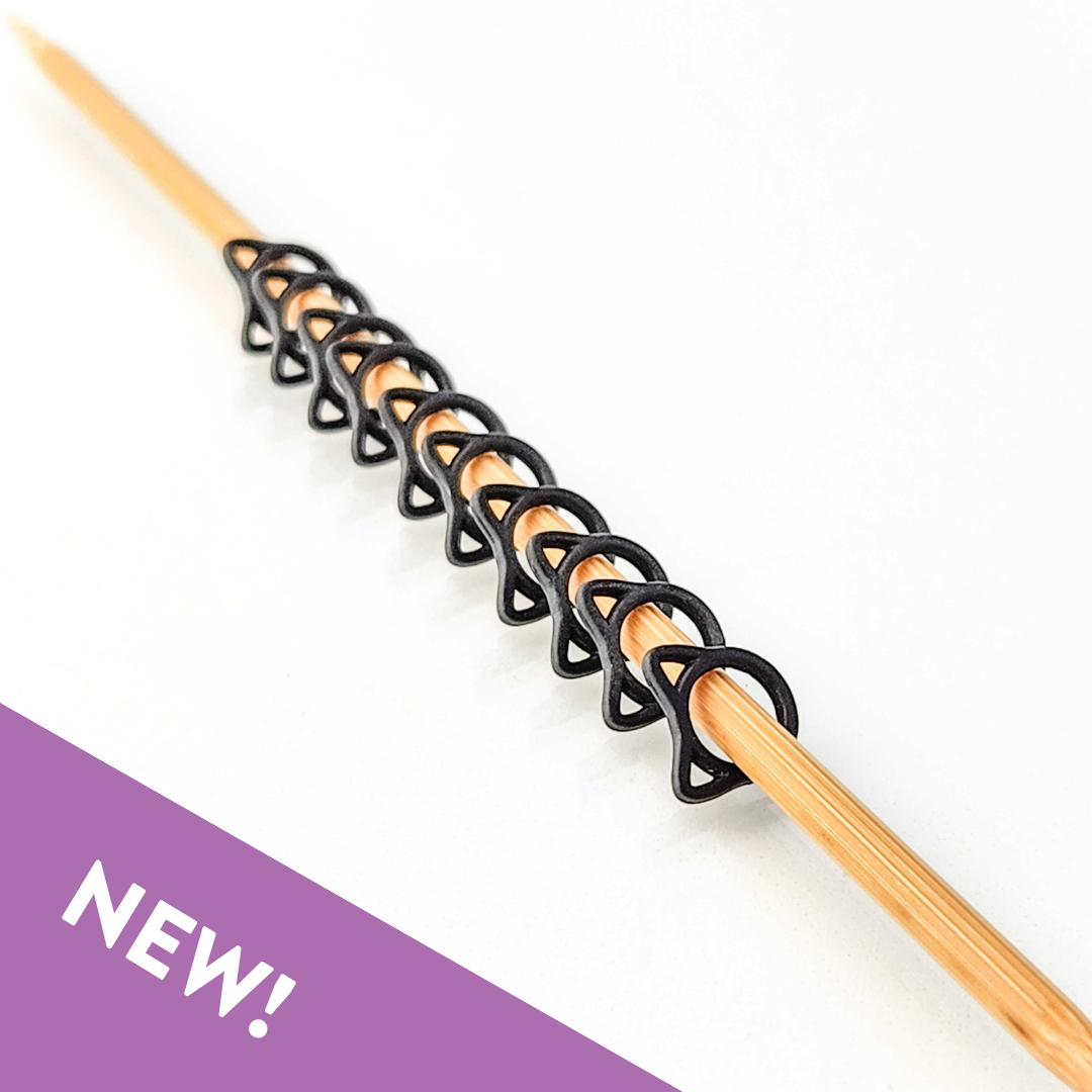 Row of black cat-shaped snag free stitch markers lined up on a bamboo knitting needle, with a purple NEW banner