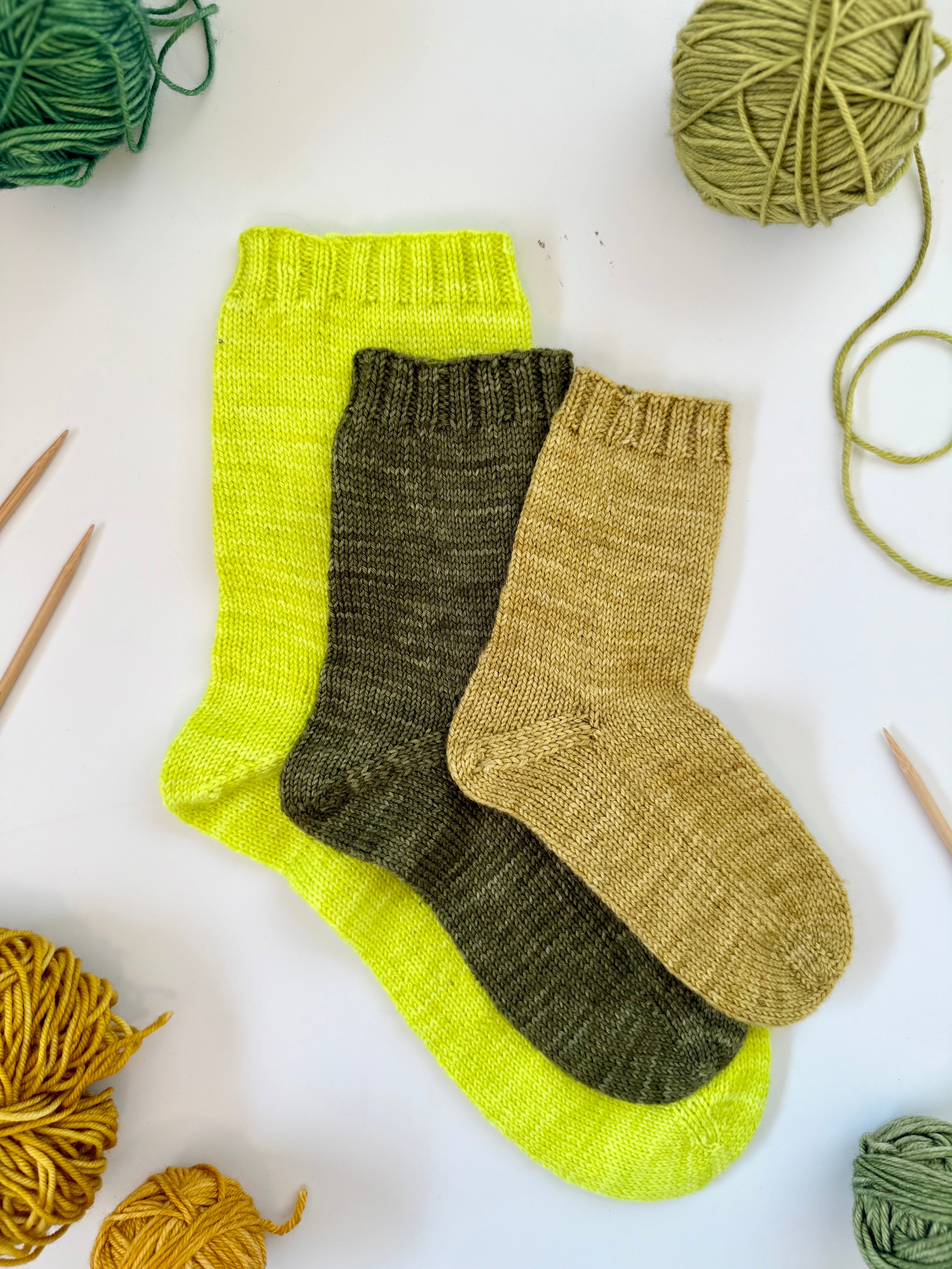 Three Base Doodle Toe-Up Socks in neon yellow, olive and gold with yarn and needles