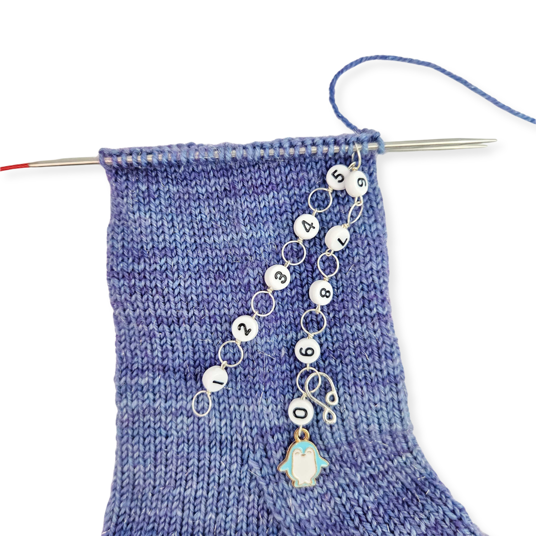 Blue stockinette knit swatch on bamboo needles with silver button stitch markers and a chain row counter attached, from the 2026 LYS Exclusive Winter Gift Box