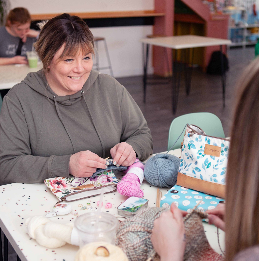 Smiling woman in an olive-gray hoodie knitting at a table with blue and pink yarn balls, a floral project bag, and a Twice Sheared Sheep leaf-print bag beside her, while another attendee knits across the table in a cafe-style event space