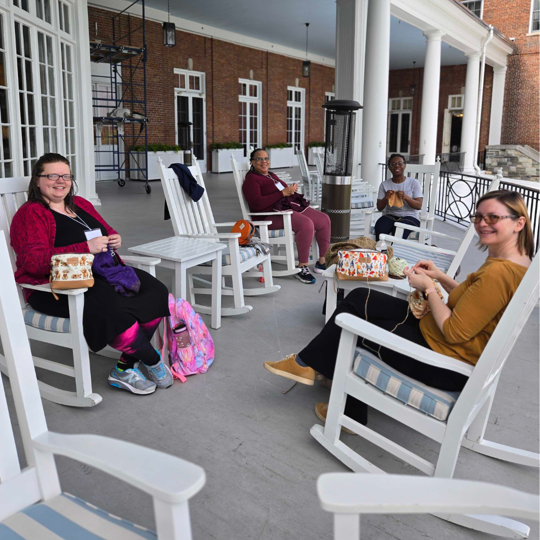 Four women sitting in white rocking chairs on the covered hotel veranda, each knitting with colorful project bags beside them, smiling at the camera. Brick building with white columns and French doors in the background, outdoor patio heater between the chairs
