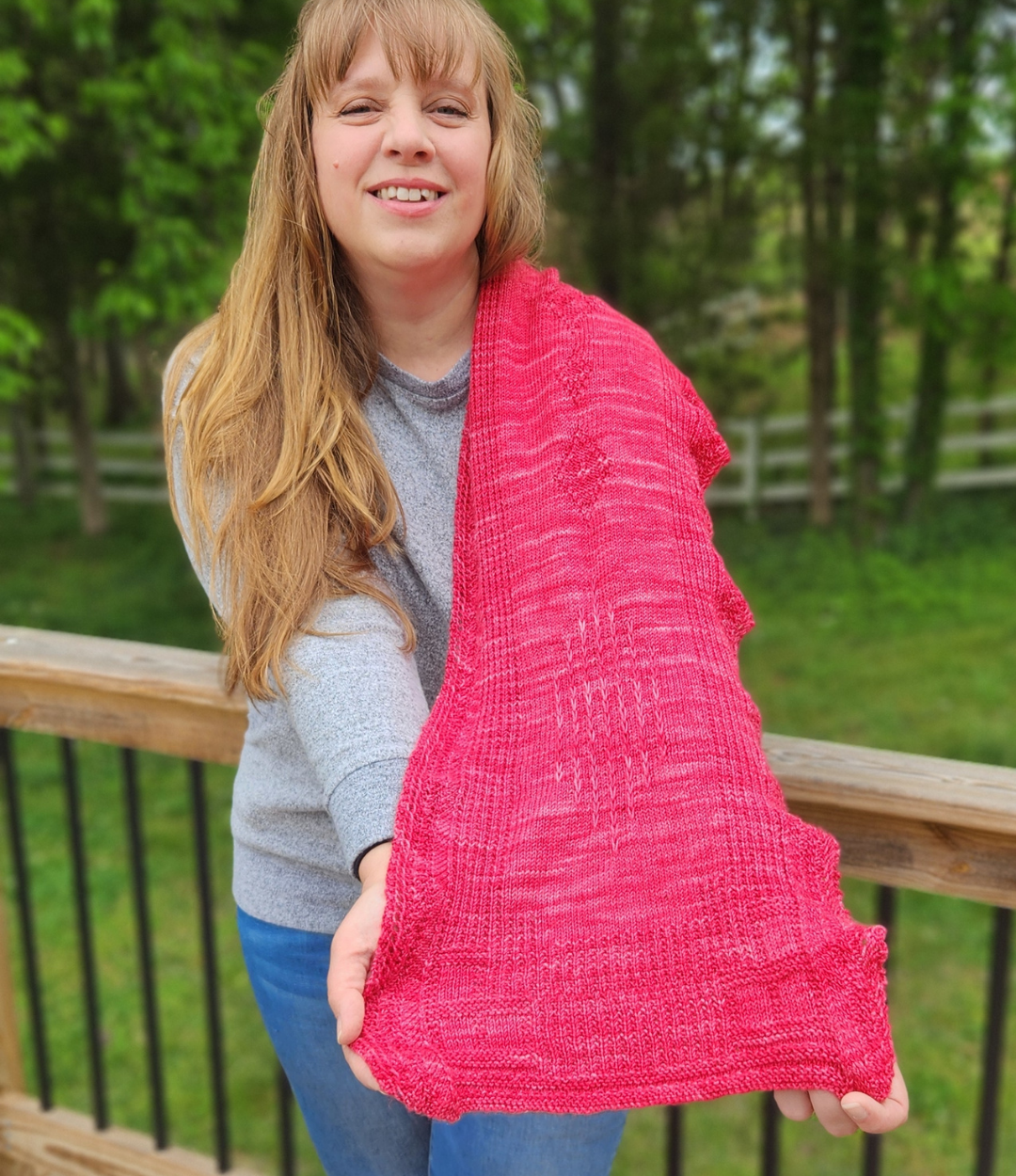 Woman holding the Revenge in Lido Wrap in coral pink lace knit fabric up to show the drape and openwork stitch pattern