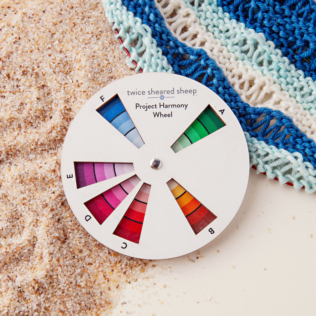Project Harmony Color Wheel resting on sand beside a blue and cream knitted shawl