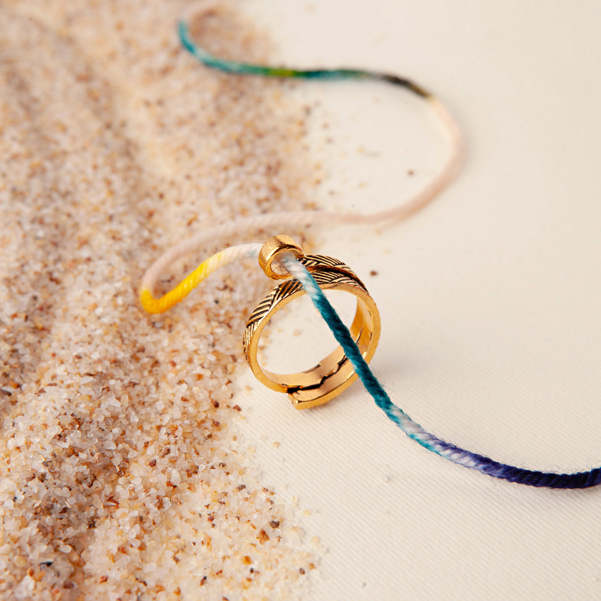 Gold feather-patterned yarn tension ring on sand with a strand of variegated yarn running through it
