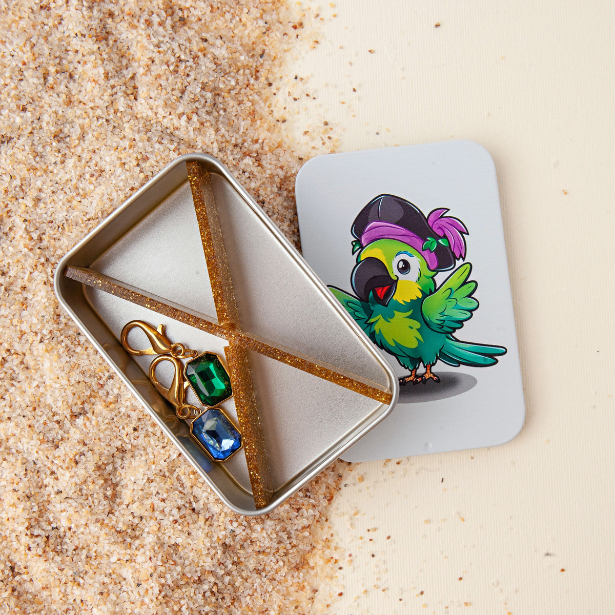 Gold glitter tin dividers crossed in an X inside an open notions tin with green and blue gem stitch markers, the pirate parrot lid beside it on sand