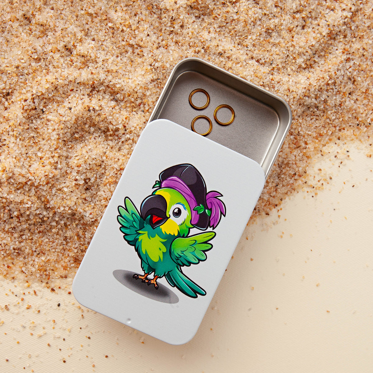 Slide-top notions tin printed with a cartoon pirate parrot, open to show three gold stitch markers, sitting on sand