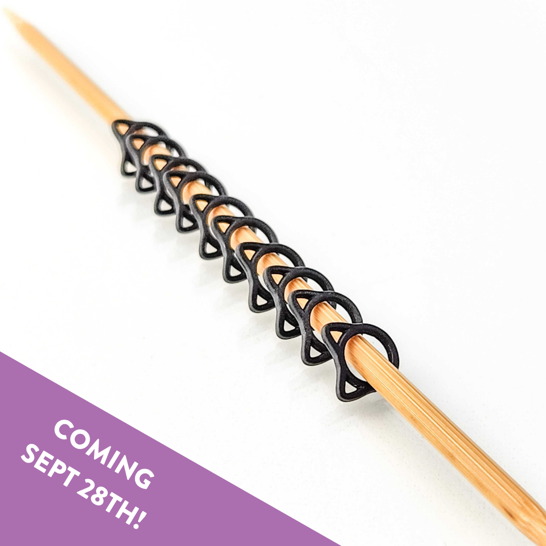 Row of black cat-shaped snag free stitch markers lined up on a bamboo knitting needle, with a Coming Sept 28th banner