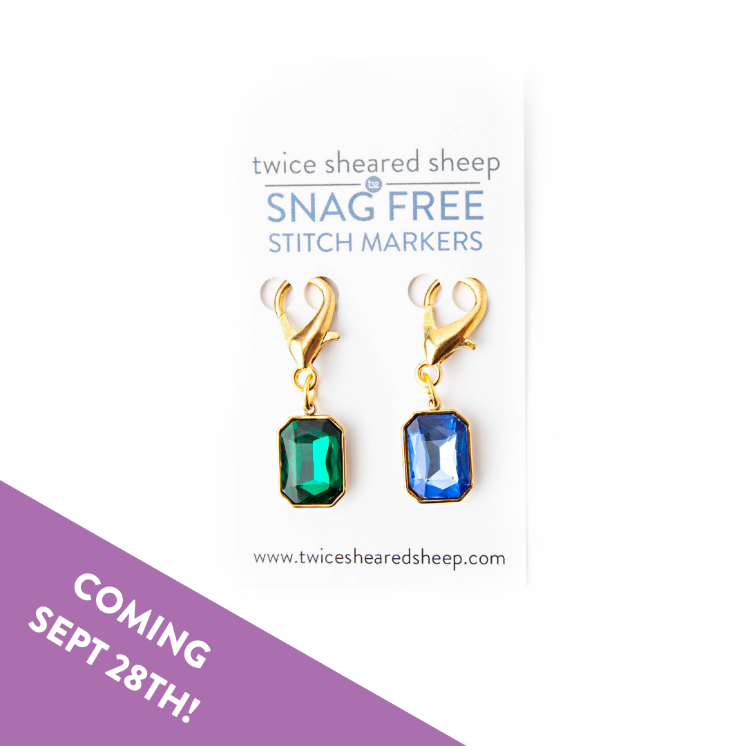 Two gold clip stitch markers with emerald green and sapphire blue jewels on a Twice Sheared Sheep Snag Free card, with a Coming Sept 28th banner