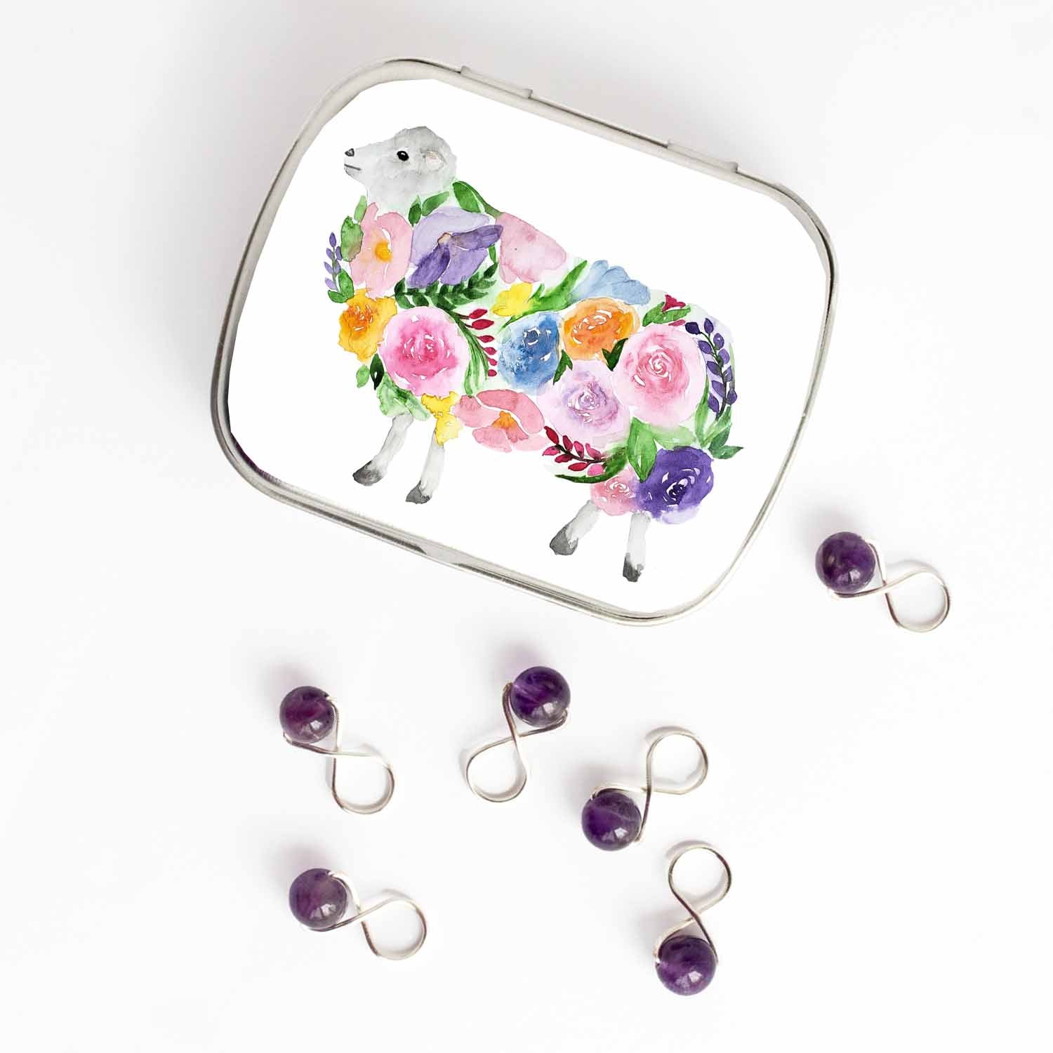 Small hinged Floral Sheep tin with watercolor flower-covered sheep lid, shown beside six purple beaded stitch markers