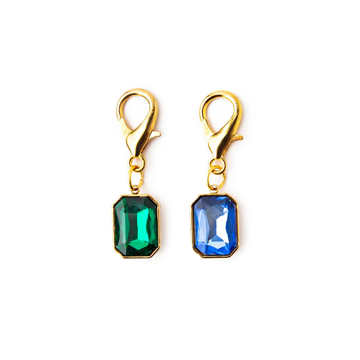 Two gold snag-free stitch markers with emerald green and sapphire blue rectangular gem charms and lobster clasps on a white background