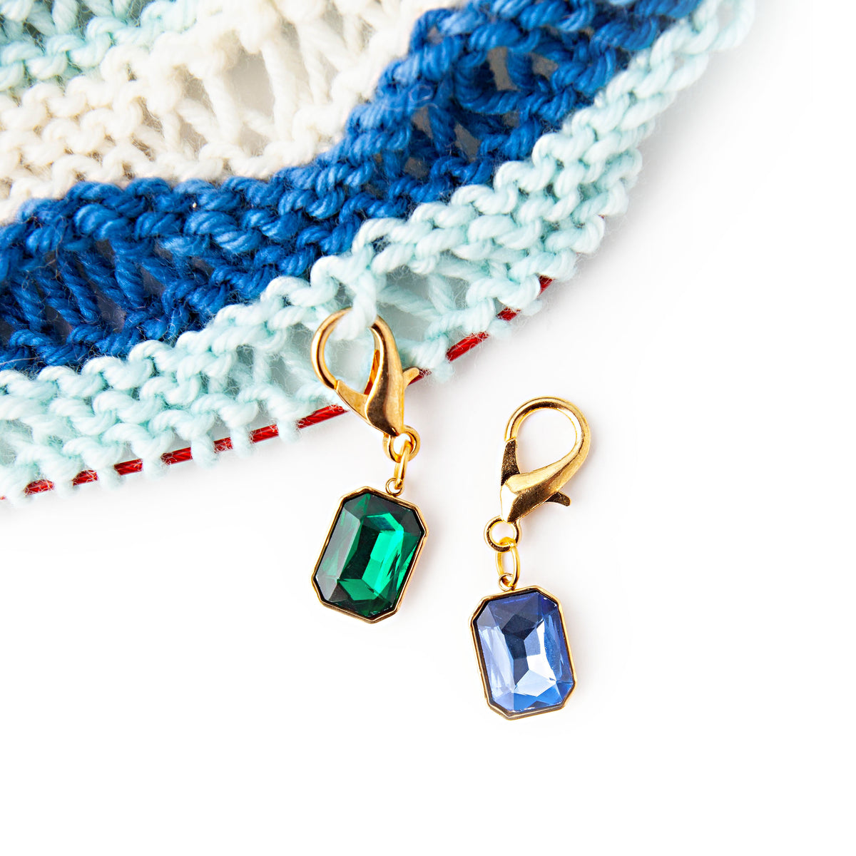 Green and blue gem stitch markers clipped to the edge of a hand-knit shawl in white, light aqua and deep blue stripes