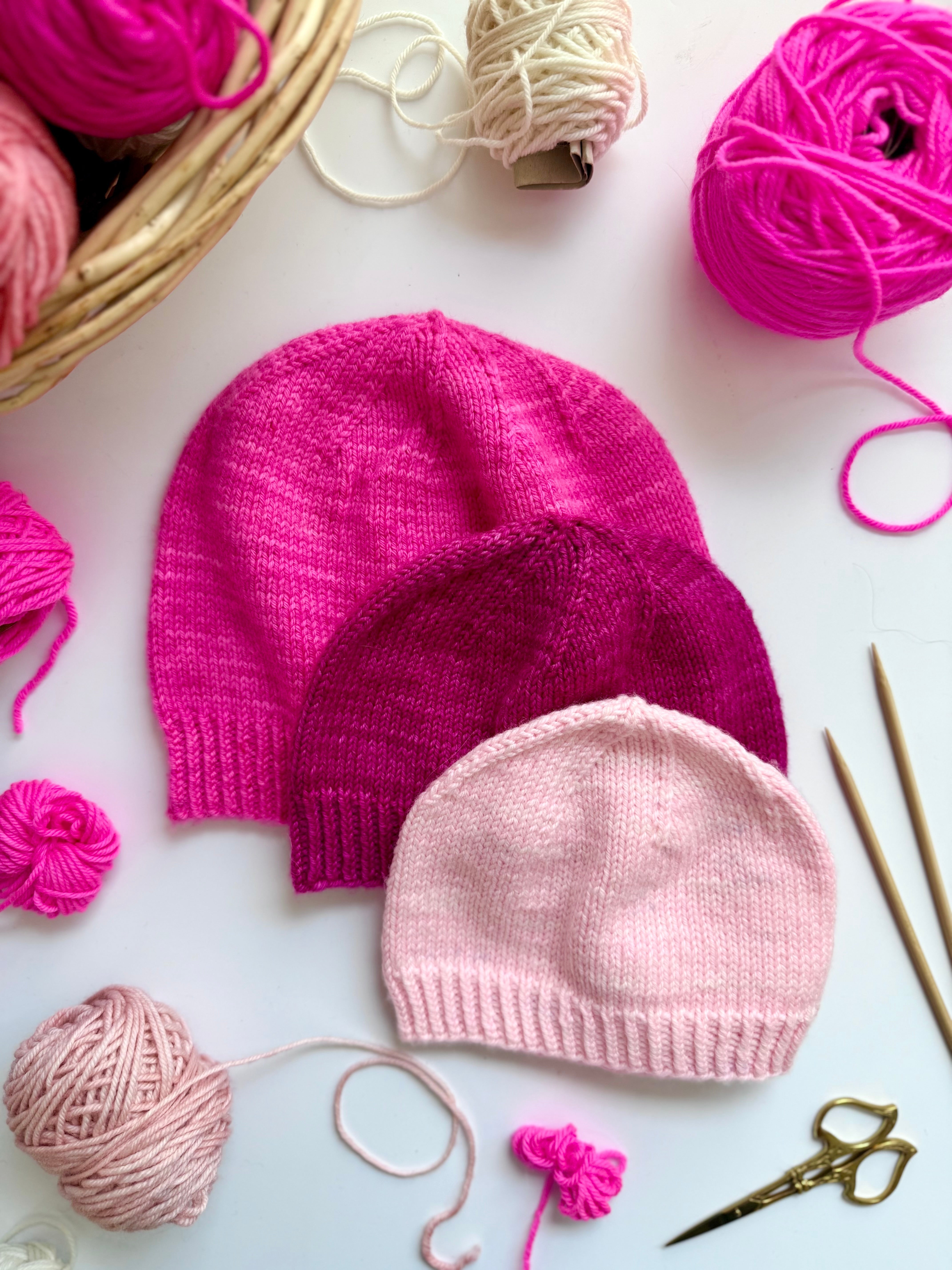 Three Base Doodle Beanie 1 hats in pink shades with yarn and needles