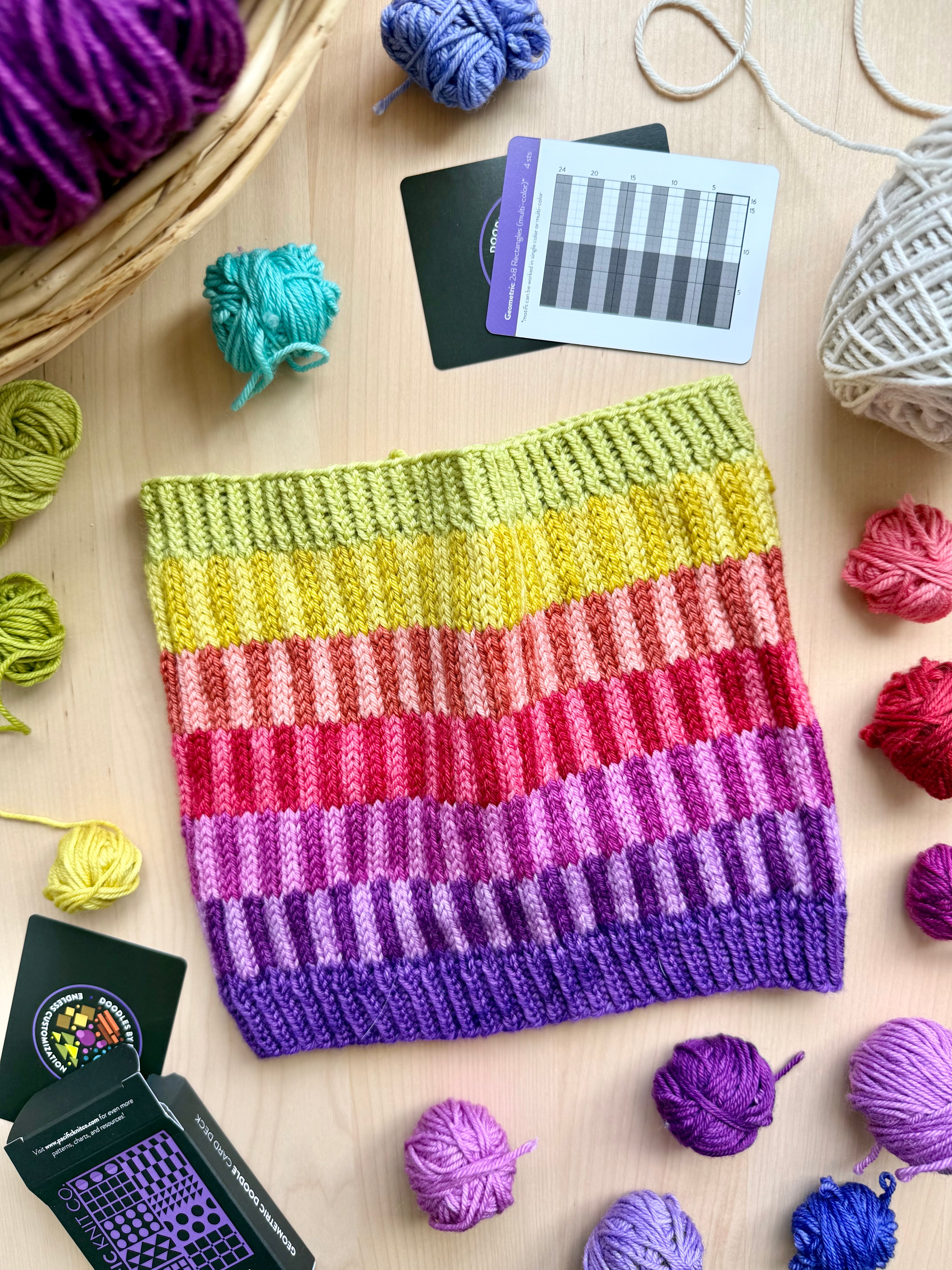Rainbow striped Geometric Doodle colorwork cowl beside the deck and chart cards