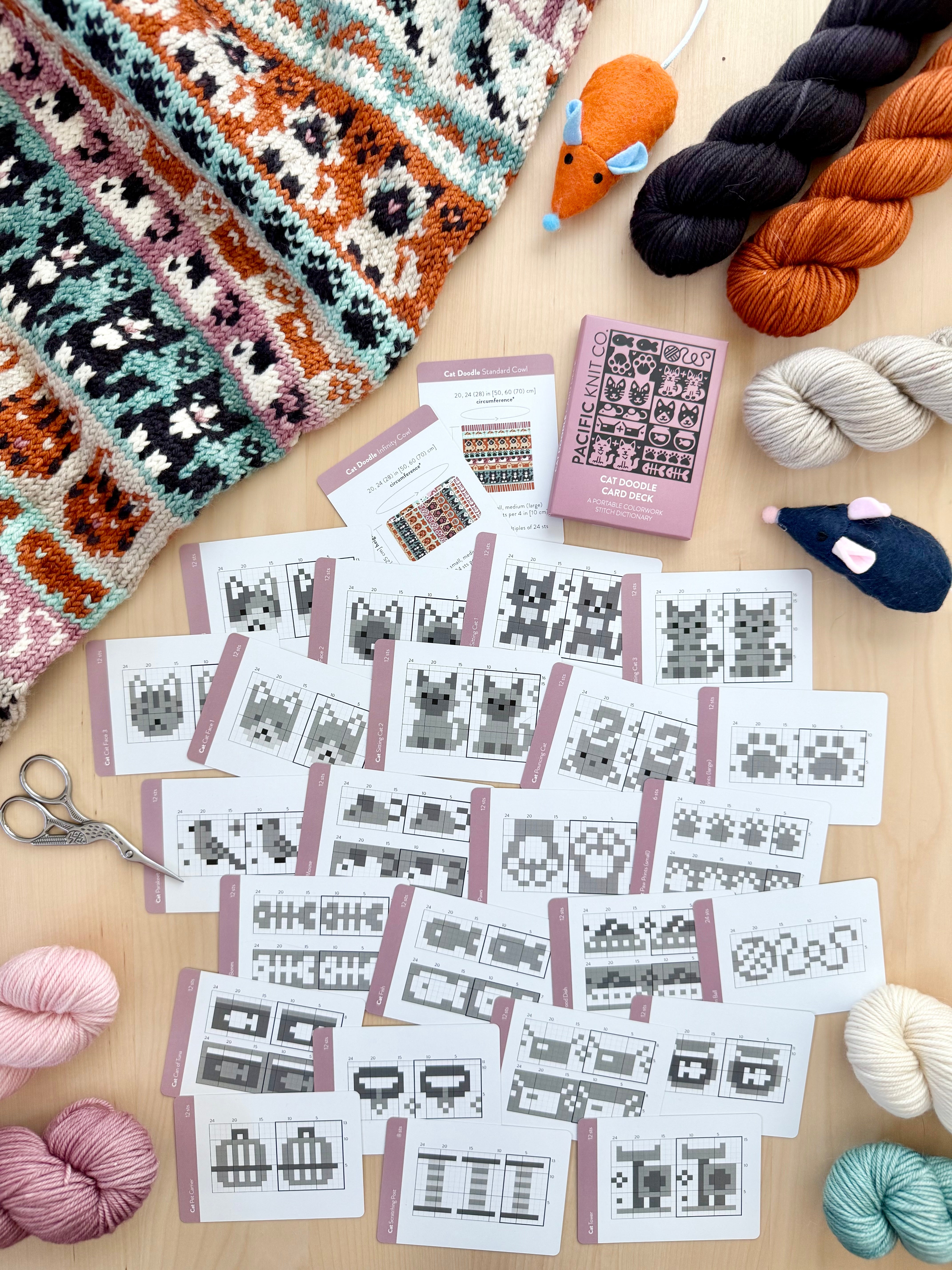Cat Doodle chart cards scattered on a wooden table with a colorwork knit and toy mice