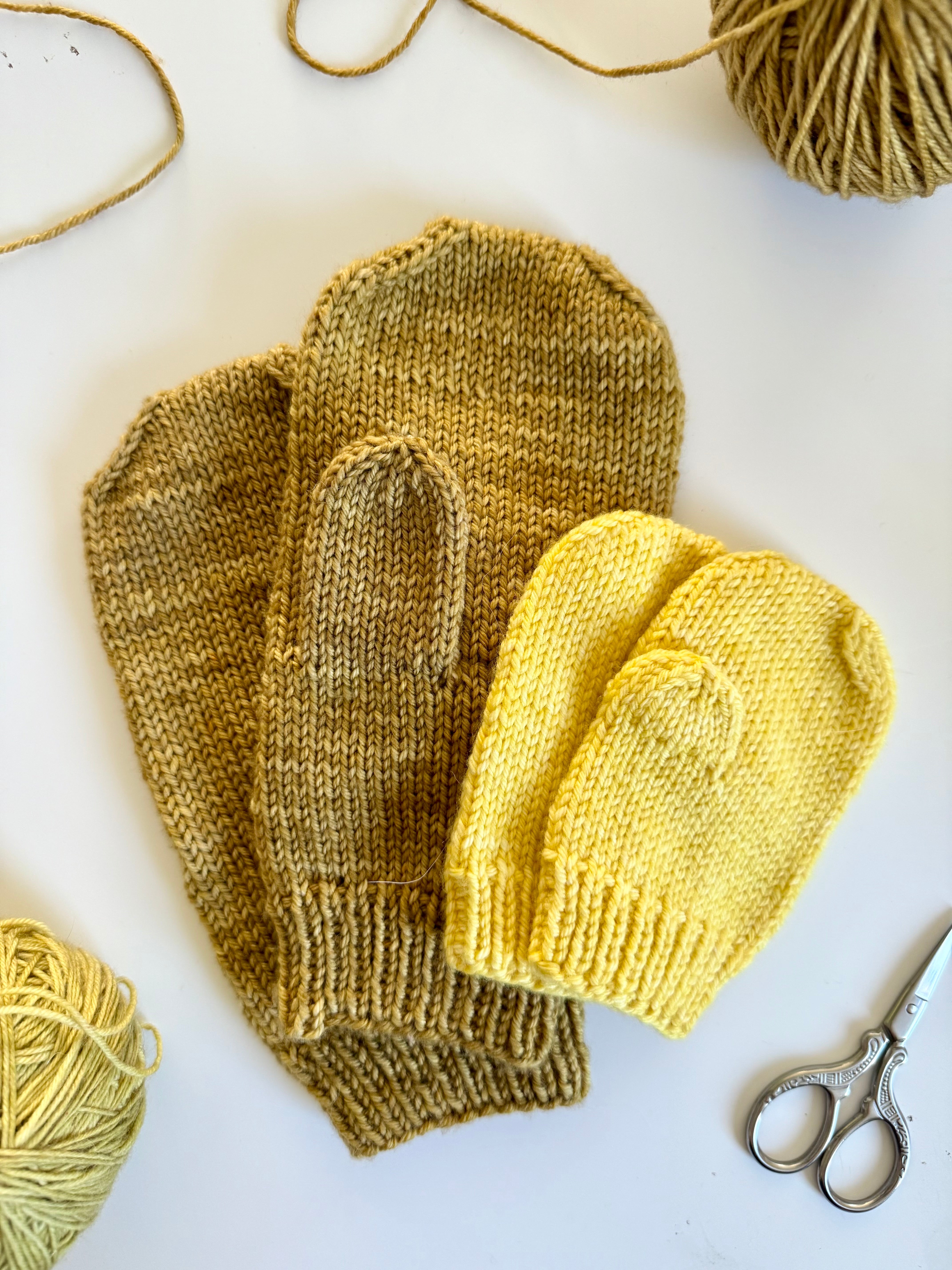 Two pairs of Base Doodle Mittens in olive and yellow with yarn and scissors