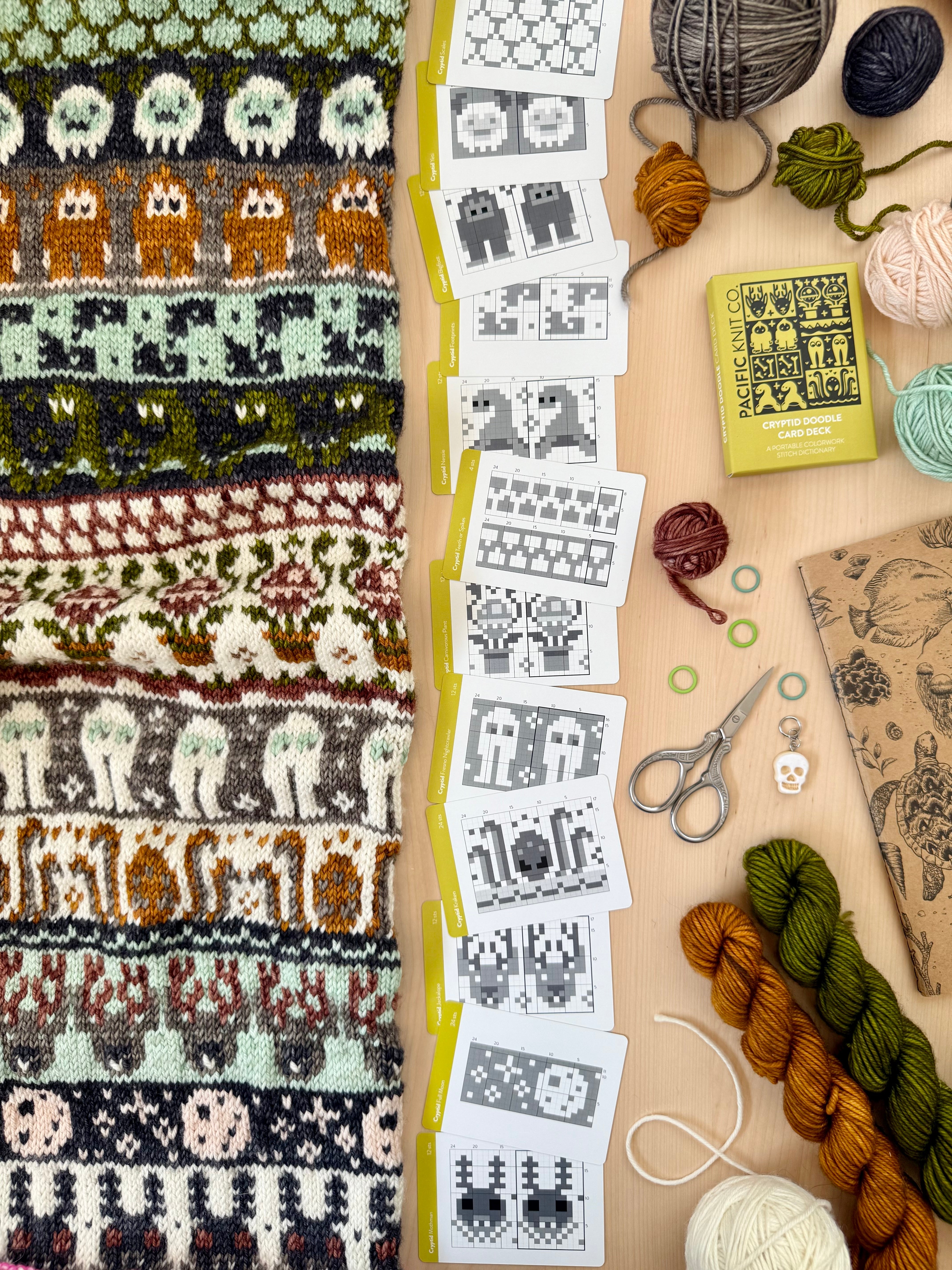Cryptid Doodle chart cards beside a monster-motif colorwork knit and mini skeins