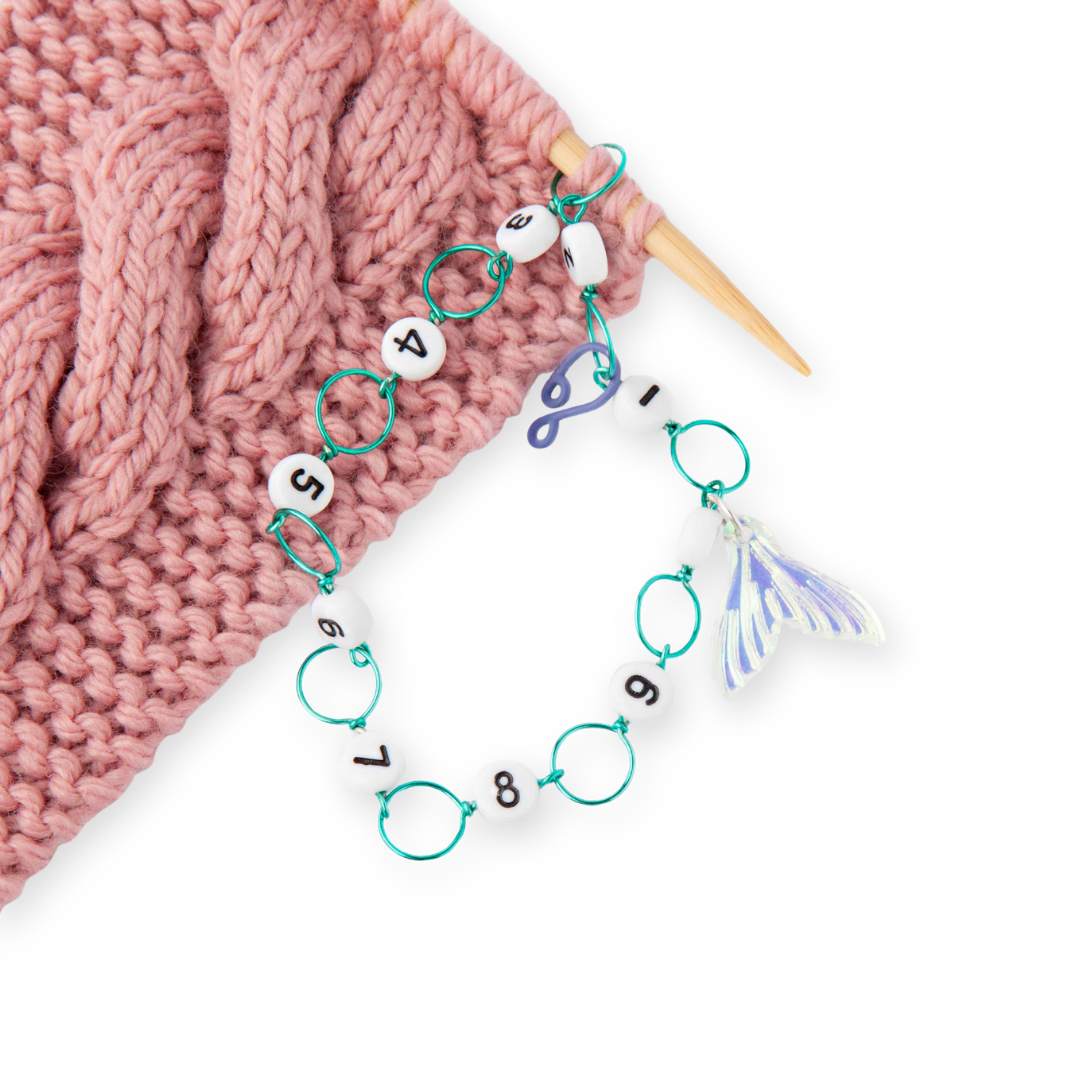 Mermaid Tail Row Counter in use on pink knitted fabric with wooden knitting needles, showing numbered beads and iridescent mermaid tail charm from the 2026 Summer Box