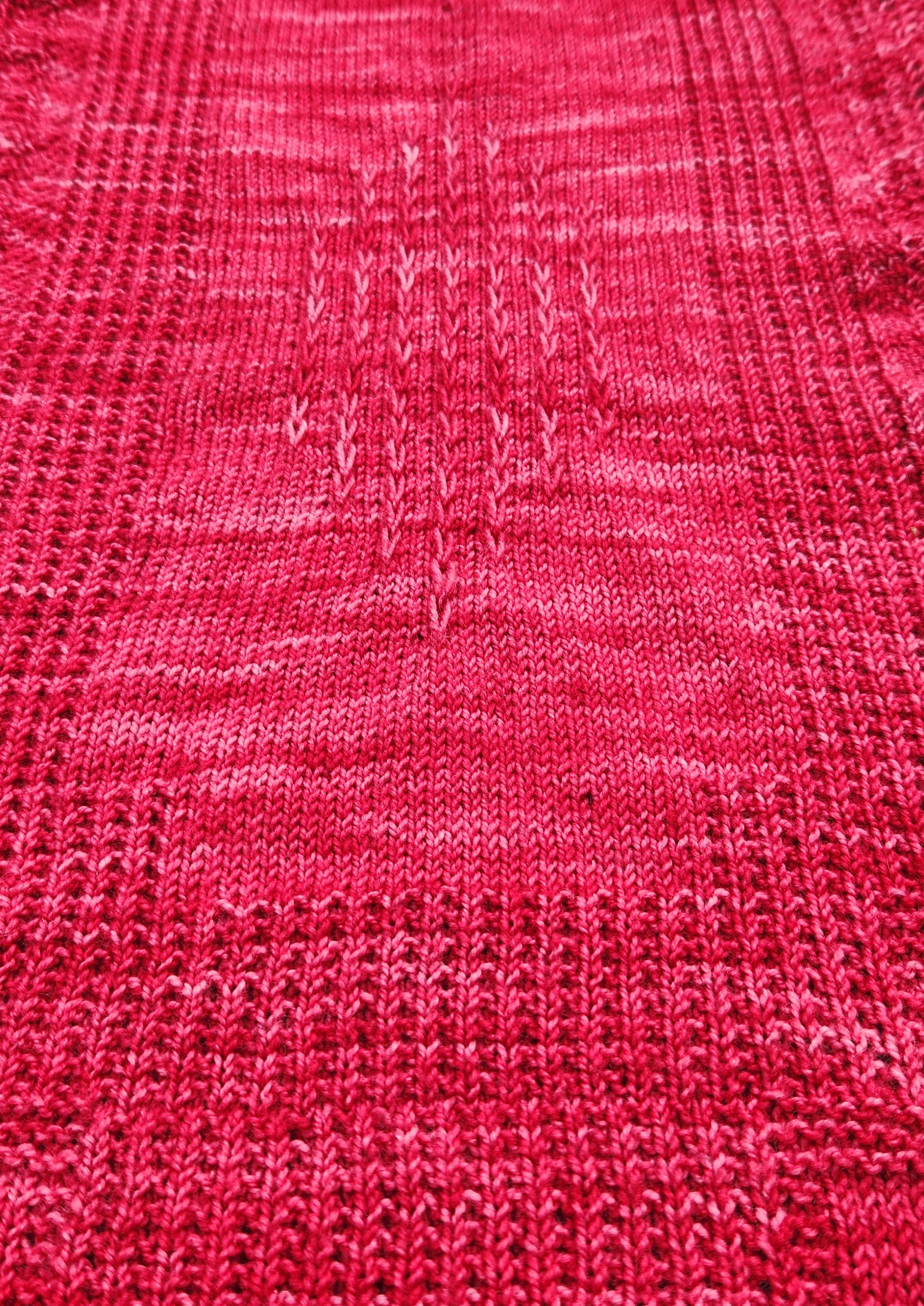 Woman holding the Revenge in Lido Wrap up flat showing the full rectangular shape and allover lace stitch pattern in coral pink fingering weight yarn