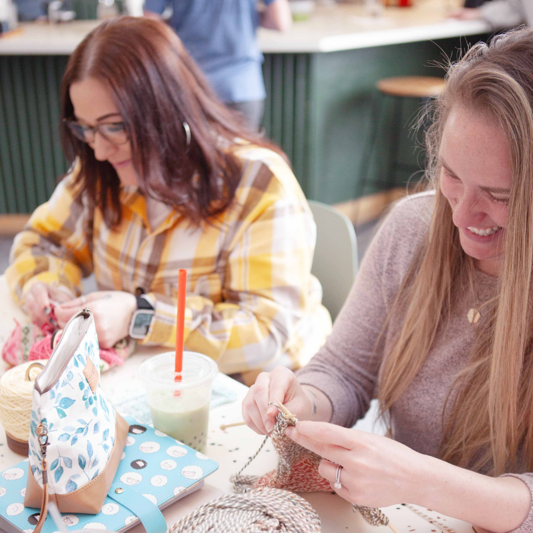 Two women knitting together at a table — one with dark hair and glasses in a yellow plaid shirt working on a pink yarn project, the other with long blonde hair in a mauve sweater smiling while knitting. Iced green drink, floral and sheep-print project bags on the table