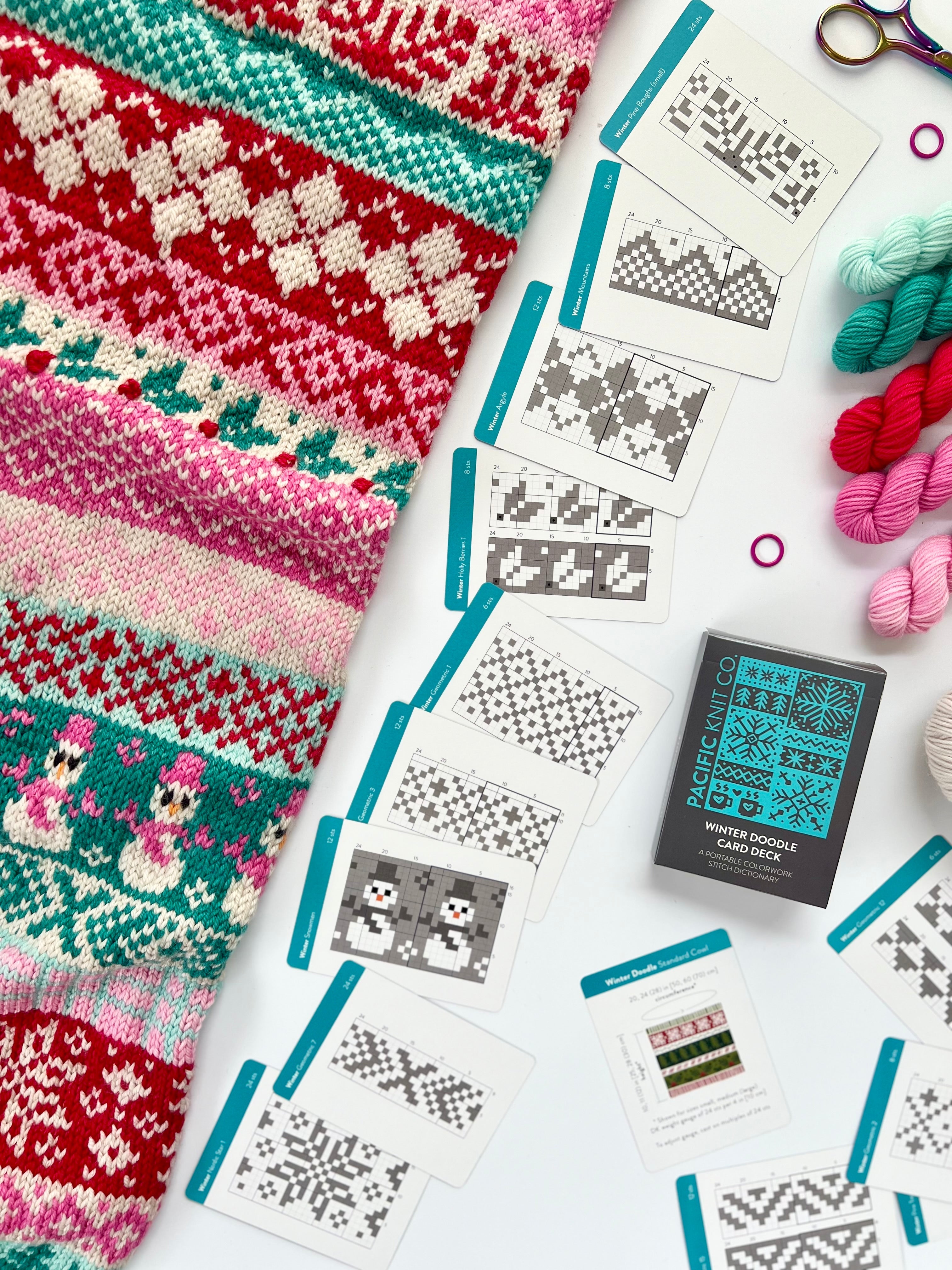 Winter Doodle chart cards fanned beside a red and green Fair Isle knit and mini skeins